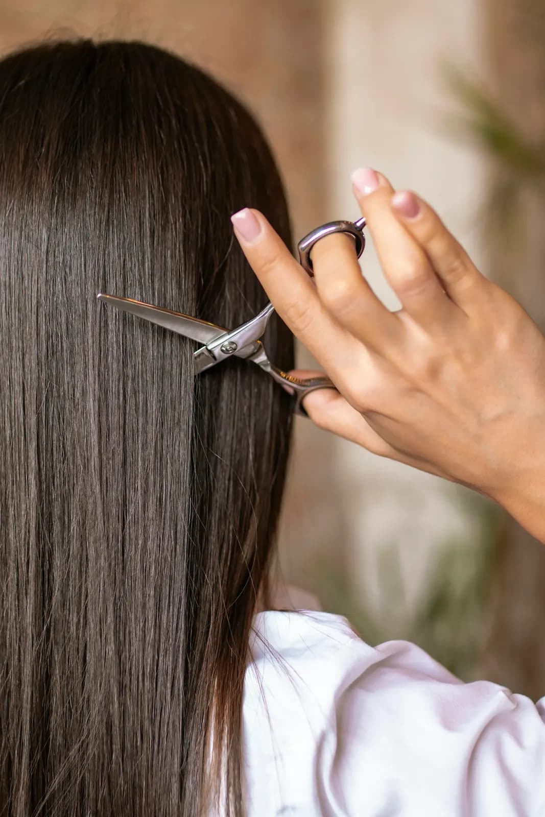 A hand with pale pink nails point-cutting long, straight dark hair with scissors