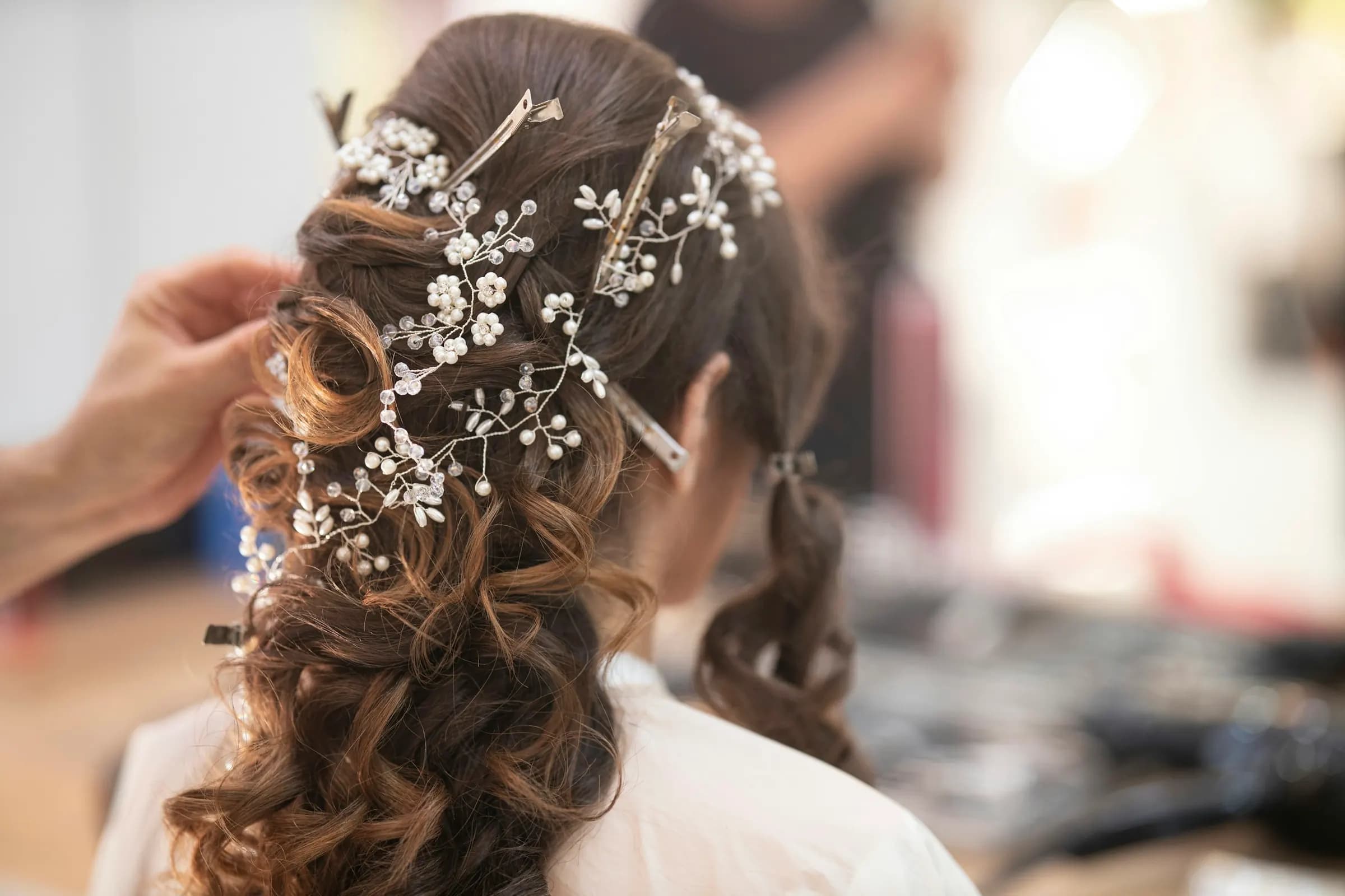 Seen from behind, a stylist's hand pins soft brown curls into a half-up style threaded with a pearl hair vine