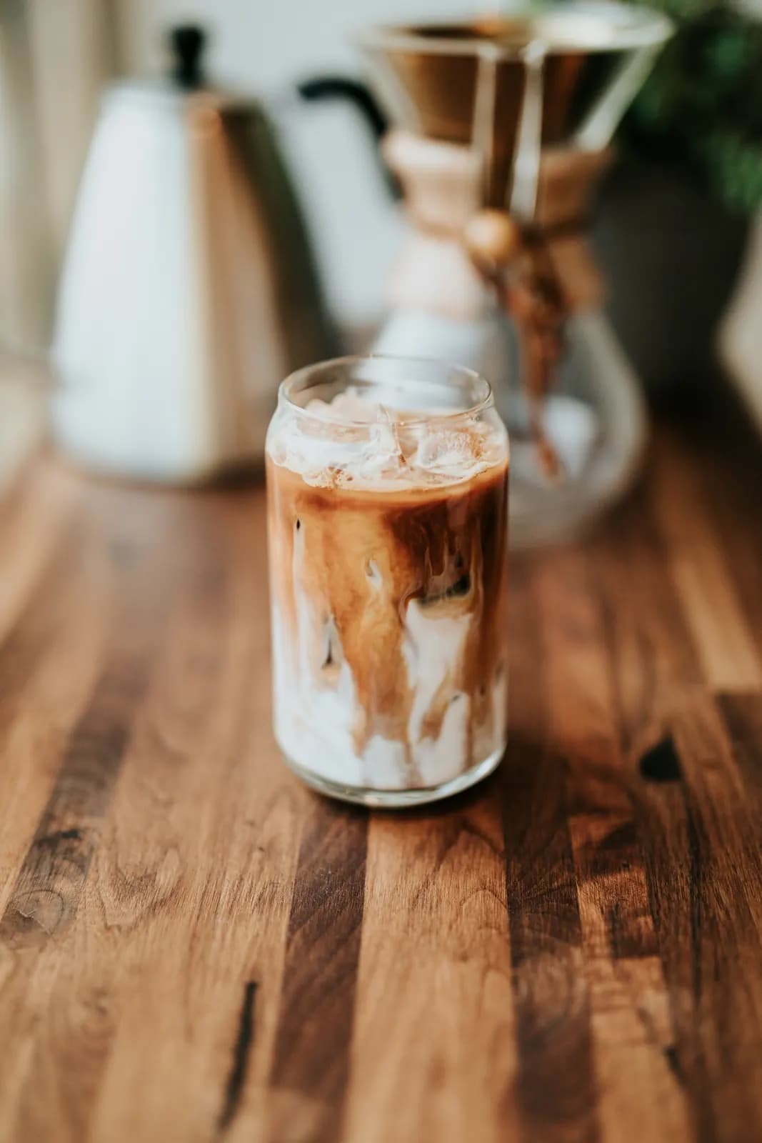 An iced latte on a wooden bar, with a Chemex and a kettle behind it