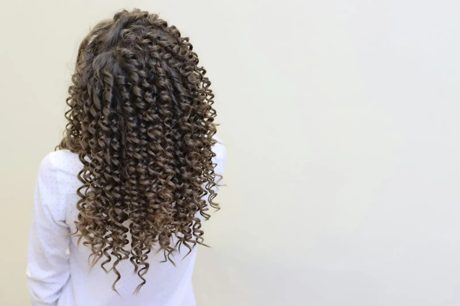 Long brown spiral curls seen from behind, against a cream wall