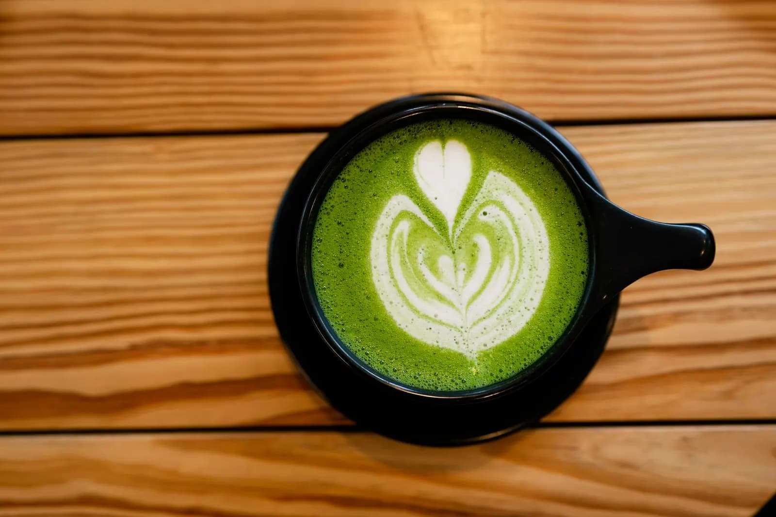 Seen from above, a matcha latte with heart art in a black cup on wooden slats