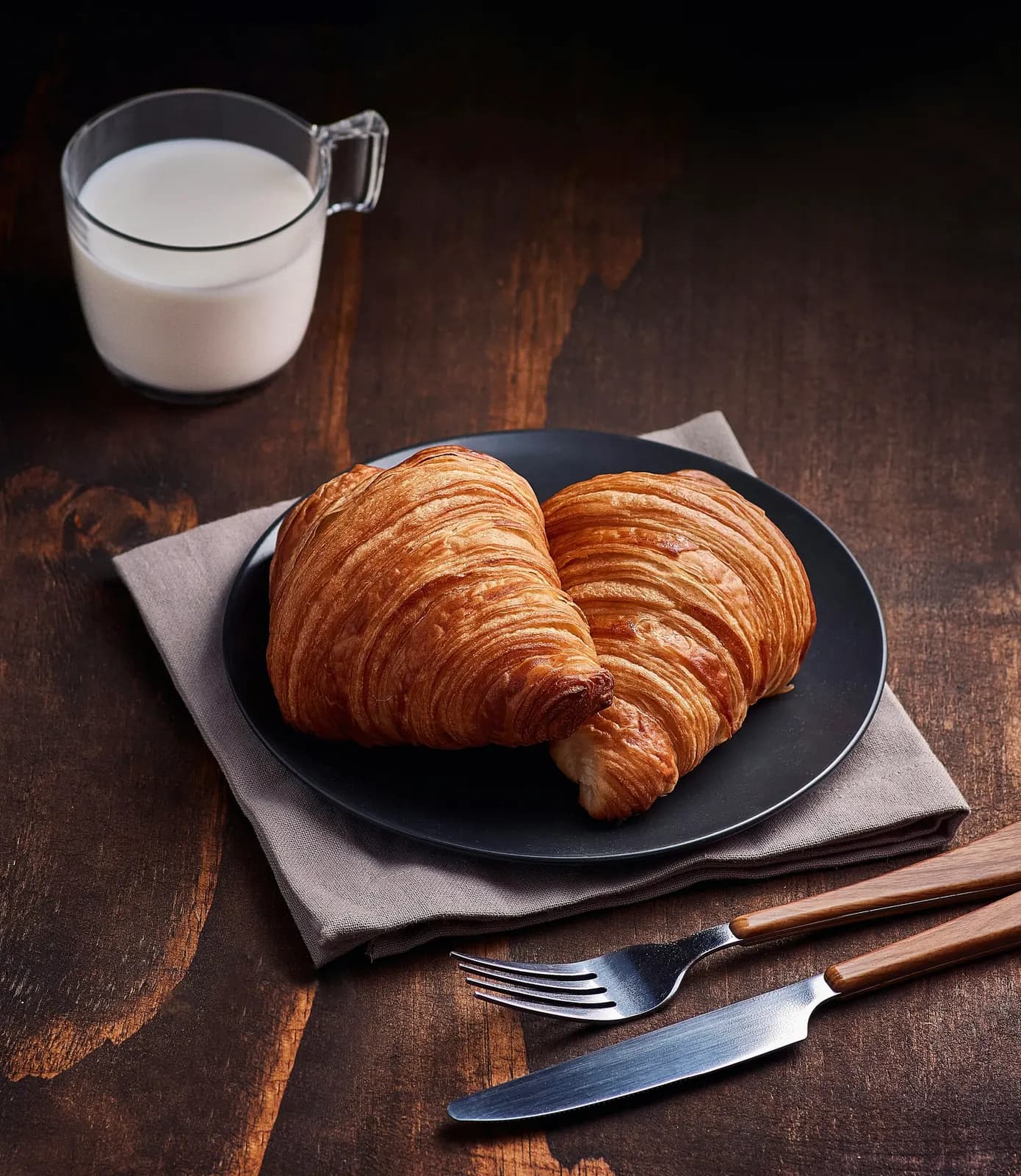 Two croissants on a dark plate beside a glass of milk