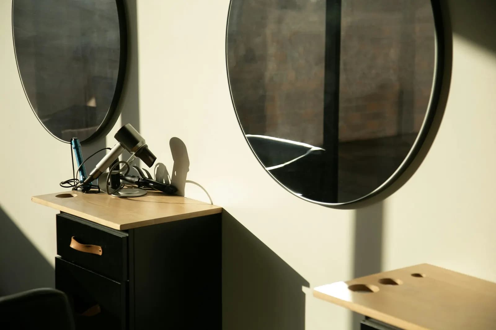 Two round mirrors above wood-topped stations, a hairdryer and curling tongs in warm sunlight