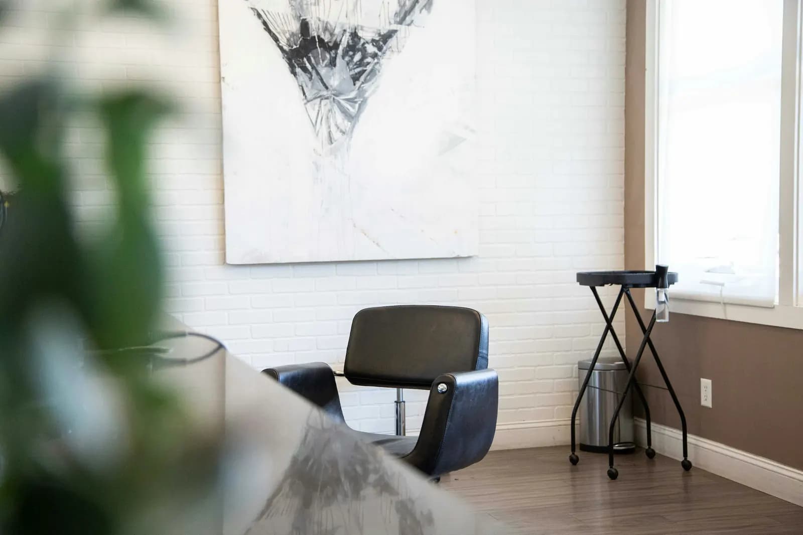 A quiet salon corner: a black styling chair by a window, under a large abstract painting