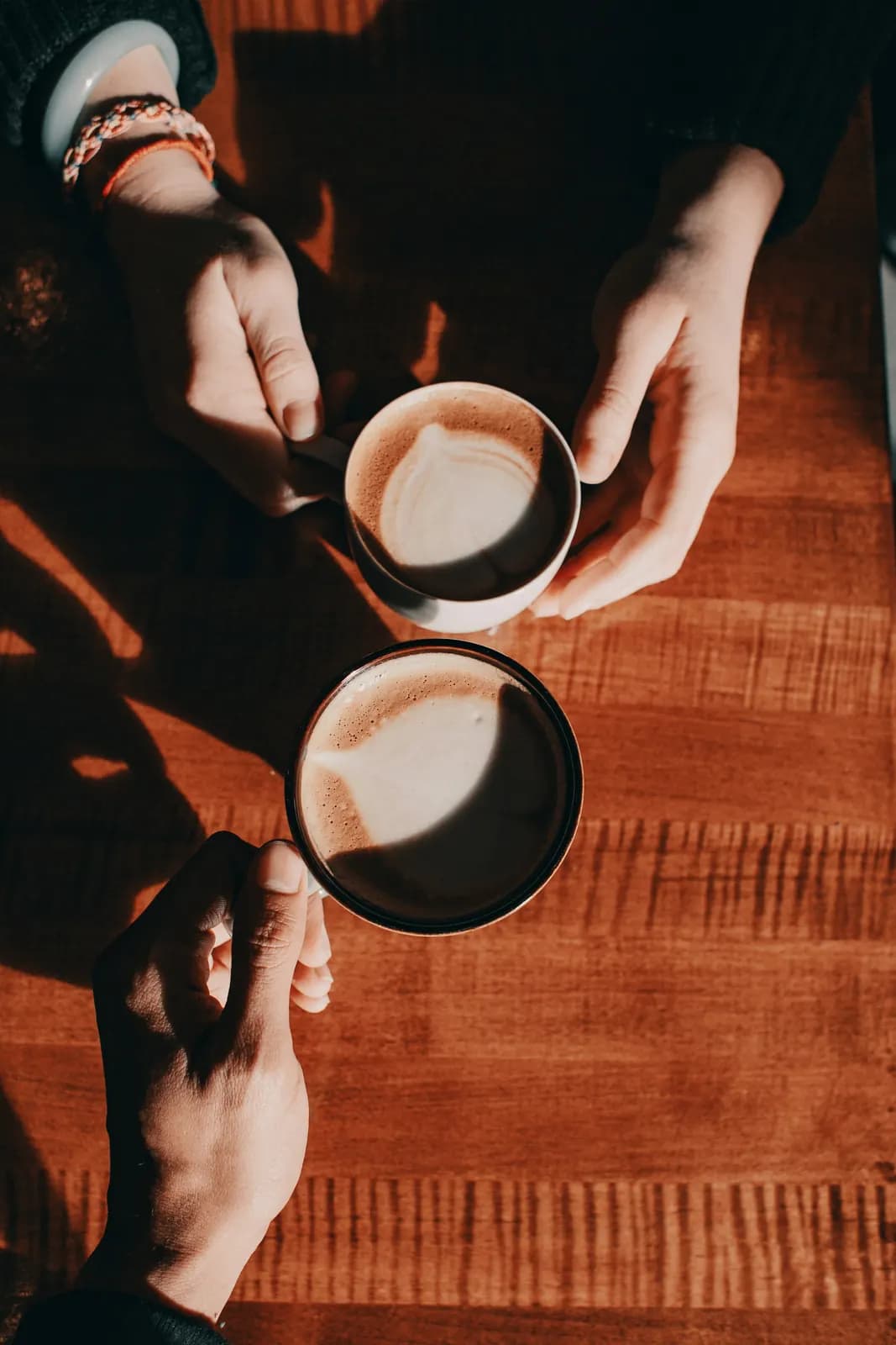Seen from above, two people holding lattes over a warm wooden table